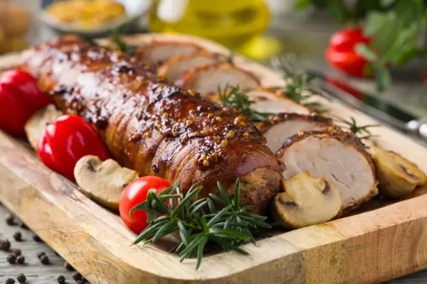 Lomo de cerdo glaseado con mostaza, servido con champiñones, tomates y romero en una tabla de madera.