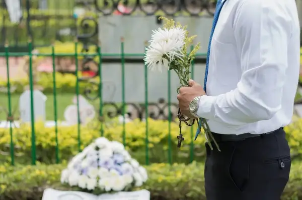 Mężczyzna w białej koszuli i niebieskim krawacie trzyma bukiet białych kwiatów i różaniec. W tle widać nagrobki i zielone krzewy. Msza pogrzebowa z urną trwa.