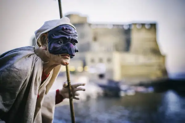 Pulcinella con maschera viola, pronto a litigare in napoletano, con Castel dell'Ovo sullo sfondo.