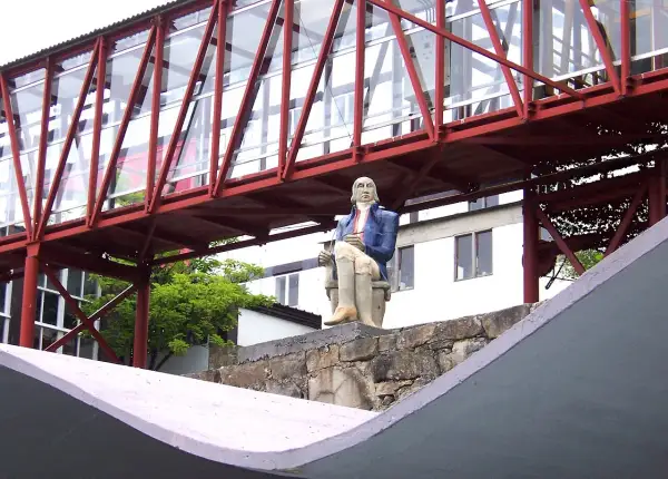 Estatua de un hombre sentado frente a la fábrica de Sargadelos, bajo un puente rojo y acristalado.