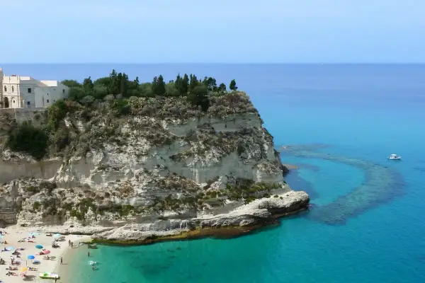 Scogliera bianca con un santuario in cima, bagnata da un mare cristallino in Calabria. Spiaggia affollata e barca all'orizzonte.