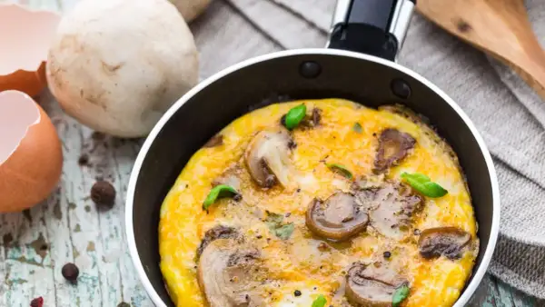 Pyszny omlet z pieczarkami, świeżo usmażony na patelni, z dodatkiem bazylii. Obok skorupki jajka i grzyby.