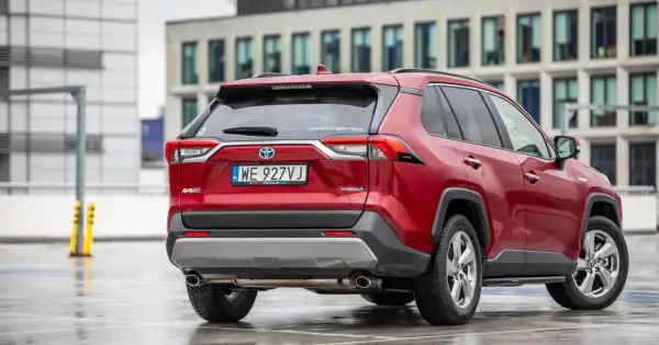 Czerwony Toyota RAV4 2020 Hybrid na mokrym parkingu, z nowoczesnym budynkiem w tle.