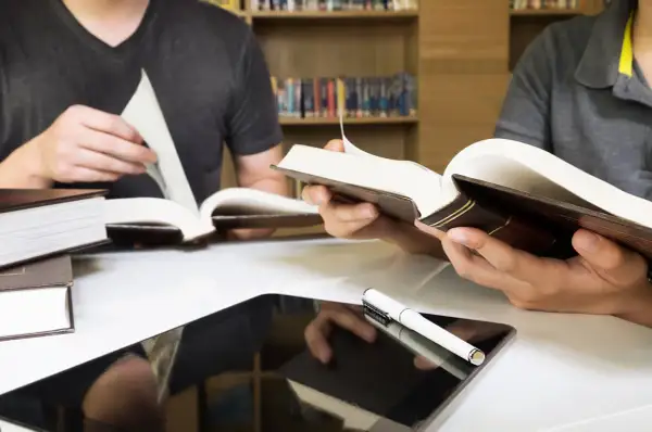 Estudiantes consultan libros y tablet en biblioteca, preparándose para las competencias específicas LOMLOE Andalucía.