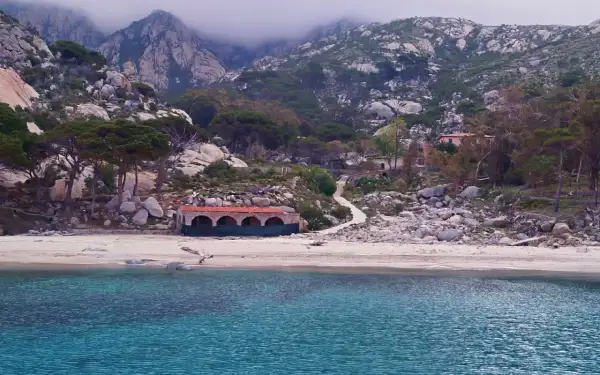 Spiaggia selvaggia con rocce e vegetazione, un edificio storico e montagne nebbiose sullo sfondo. Qui si trova l'isola di Montecristo.