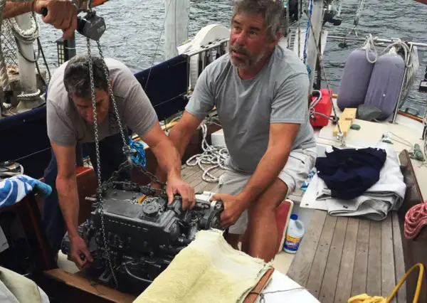Due uomini lavorano alla manutenzione del motore di una barca a vela, sollevandolo con una catena.