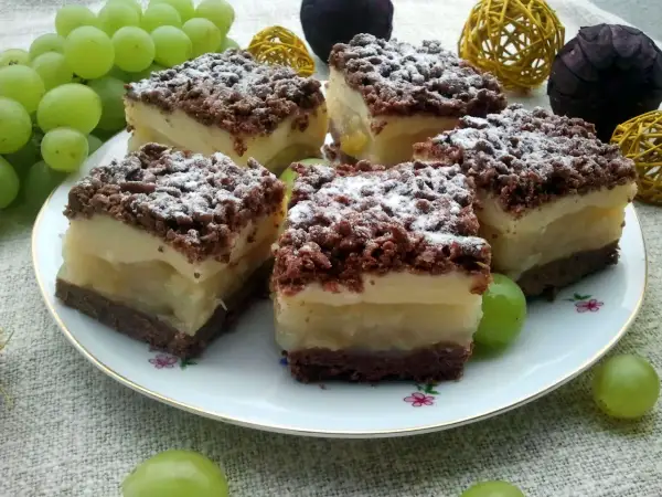Szybkie ciasto z gruszkami i budyniem, posypane cukrem pudrem, na talerzu z winogronami.