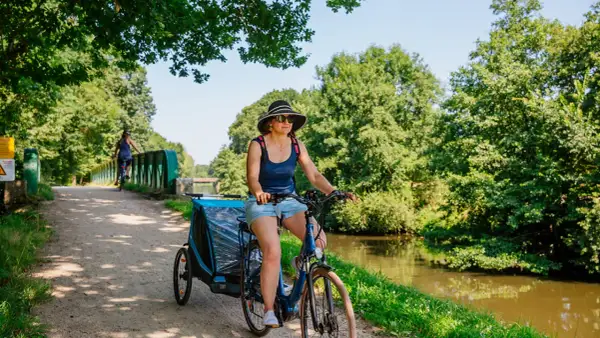 Mujer pedalea por las vías verdes de Francia con un remolque para niños, disfrutando del paisaje junto al canal.
