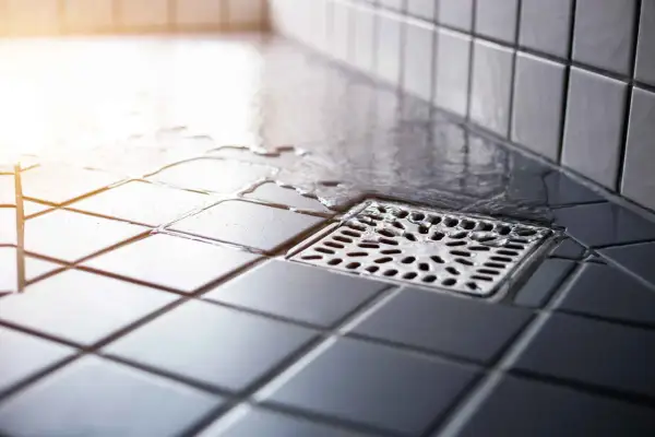 Acqua stagnante vicino allo scarico doccia puzza. Le piastrelle scure e bagnate creano un'atmosfera poco invitante.