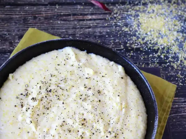 Krämig italiensk polenta i en gjutjärnspanna, strödd med svartpeppar. Majsgryn syns på den mörka träytan.