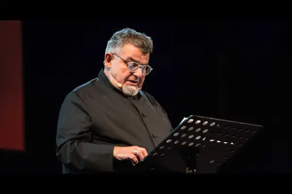 Giancarlo Cauteruccio, con occhiali e barba, legge da un leggio su un palco buio.