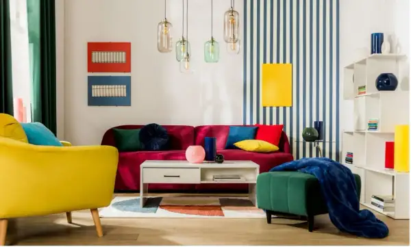 Salon w stylu memphis: żółty fotel, bordowa kanapa, kolorowe poduszki, geometryczny dywan i lampy.