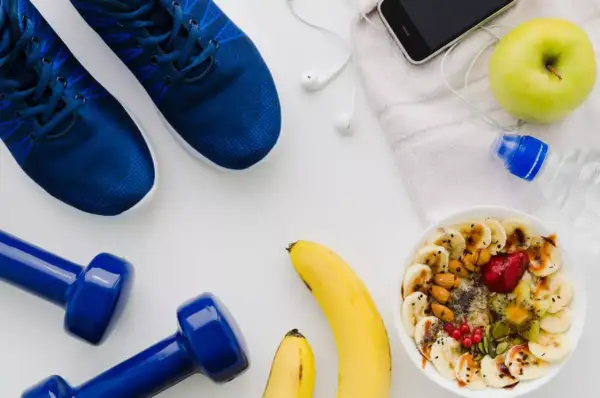 Niebieskie buty sportowe, hantle, miska owsianki z owocami, jabłko, telefon i butelka wody.