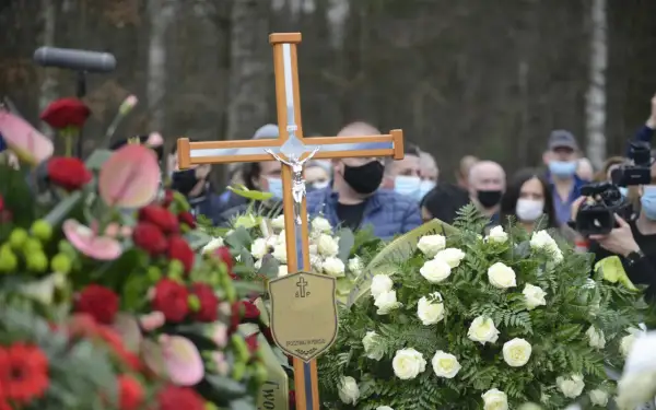Drewniany krzyż z Jezusem, otoczony wieńcami, symbolizuje pożegnanie. Po pogrzebie tabliczka z napisem "Spoczywaj w pokoju" pozostaje jako pamiątka.