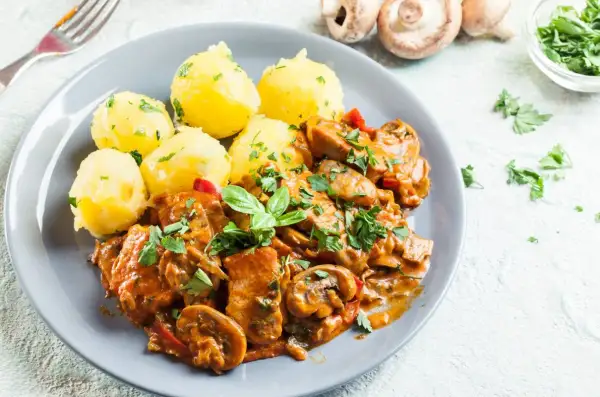 Pyszny gulasz z kurczaka z pieczarkami i papryką, podany z ziemniakami posypanymi natką pietruszki.