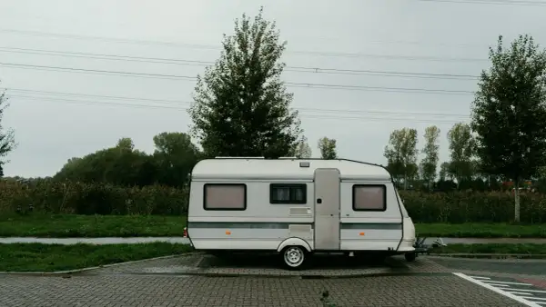 Ein weißer b96 wohnwagen steht auf einem gepflasterten Parkplatz, umgeben von grüner Wiese und Bäumen unter einem grauen Himmel.