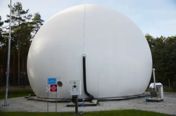 Ogromny, biały balon magazynujący biometan, otoczony drzewami. Obok znajduje się panel sterowania i rury.