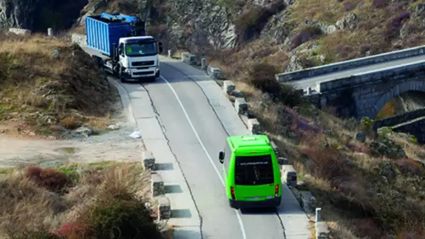 Camión blanco con contenedor azul subiendo una pendiente, seguido por un autobús verde. Se debate quién tiene prioridad en una pendiente.