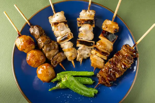 Różnorodne yakitori na niebieskim talerzu: kurczak, wątróbka, kulki mięsne i papryczki shishito.
