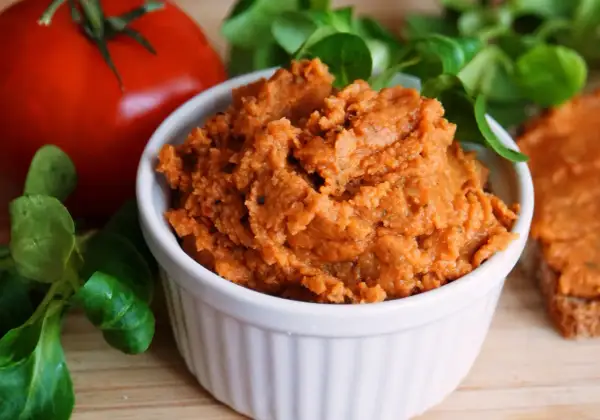 Pyszna, domowa pasta z soczewicy w białej miseczce, obok pomidora i świeżej zieleniny. Idealna na kanapki.