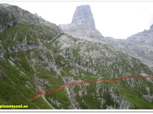 Sendero marcado en rojo asciende por la ladera verde hacia el imponente Sotres Urriellu, una cumbre rocosa en los Picos de Europa.