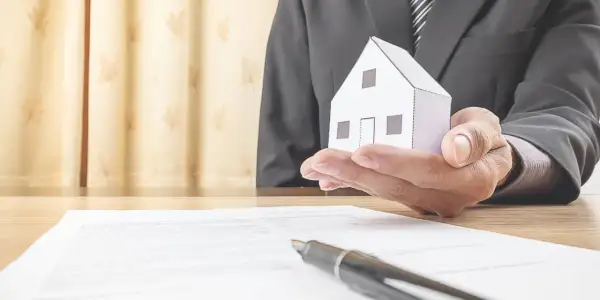 Un uomo in giacca e cravatta tiene una casetta di carta, simbolo di un accordo di leasing immobiliare per privati.