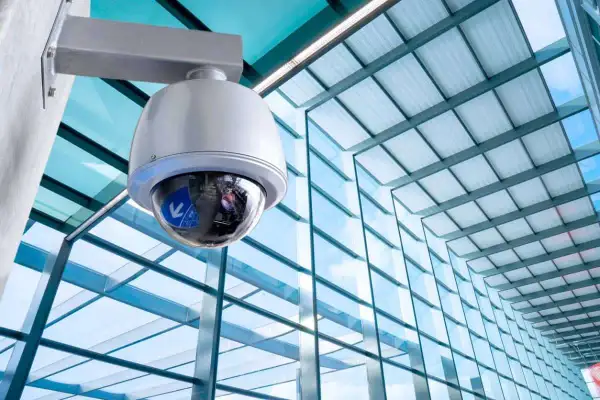 Caméra de surveillance fixée à un mur, prête à filmer. Le bâtiment moderne avec sa façade vitrée reflète le ciel bleu.