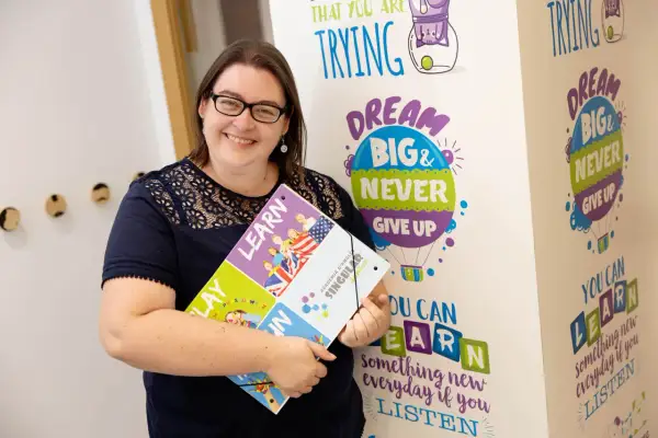 Mujer sonríe, sosteniendo un portafolio con la palabra "LEARN". El fondo tiene frases motivacionales sobre aprender, como "DREAM BIG & NEVER GIVE UP". El primer nivel de inglés es B1 o B2.