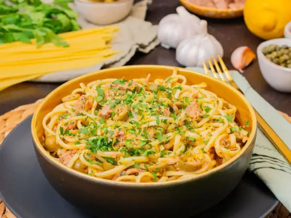 Eine Schüssel mit Thunfisch Pasta, garniert mit Petersilie und Zitronenschale. Daneben liegen Spaghetti, Knoblauch und Kapern.