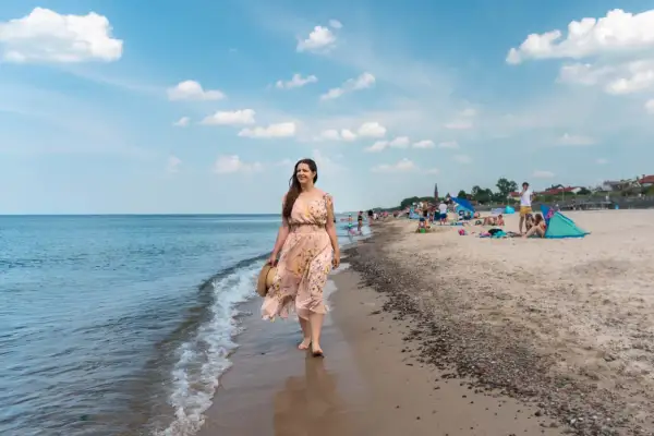 Kobieta w kwiecistej sukience spaceruje brzegiem morza. W tle plaża pełna ludzi, parawanów i namiotów – typowe sarbinowo atrakcje.