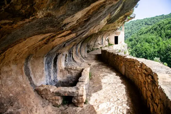 Sentiero roccioso conduce alla chiesetta di San Bartolomeo in Legio, incastonata nella roccia con vista su boschi rigogliosi.
