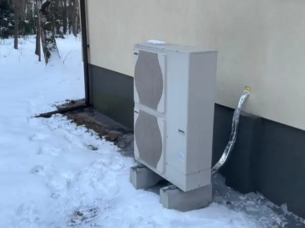 Jednostka zewnętrzna pompy ciepła stoi na betonowych podstawach w zasypanym śniegiem ogrodzie. Czy pompa ciepła jest głośna? Zimą, gdy wszystko wokół jest ciche, można to sprawdzić.