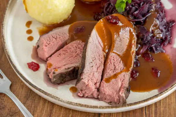 Saftiges Rehschulter Rezept: Rosa gebratenes Fleisch mit Sauce, Rotkohl und Klößen auf einem Teller.