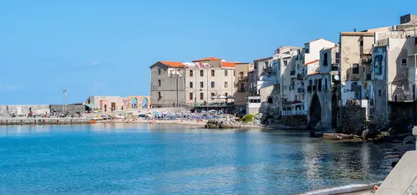 Edificios antiguos y una playa animada en Cefalú, Italia, con el mar azul brillante.