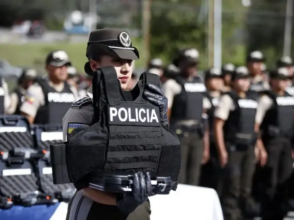 Agente de policía sostiene chaleco antibalas de material kevlar. Detrás, otros agentes uniformados.