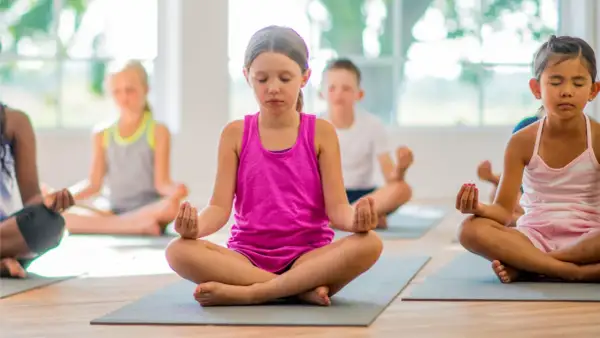 Niños practicando ejercicios de respiración y meditación en un salón de clases, sentados en colchonetas con los ojos cerrados.