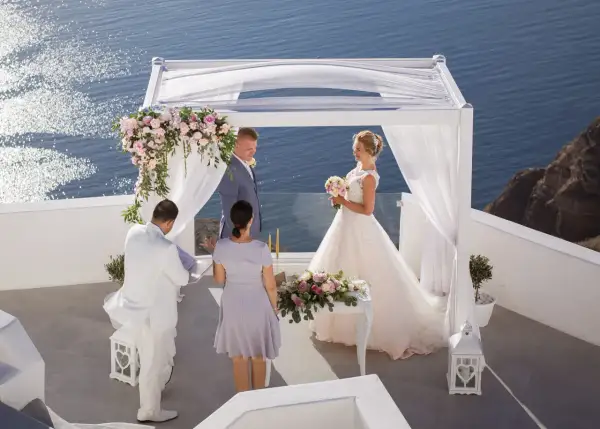 Piękny ślub w Grecji. Para młoda stoi pod białym baldachimem, w tle błękitne morze. Ceremonia odbywa się na tarasie z widokiem na ocean.