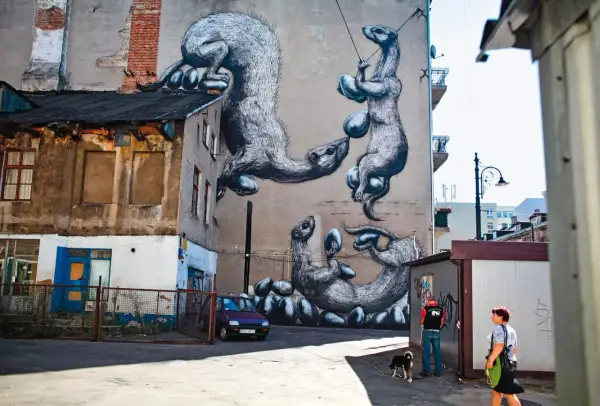 Niesamowite street art przykłady: wielkie, futrzaste zwierzęta bawią się jajkami na ścianie kamienicy. Obok przechodzą ludzie z psem.