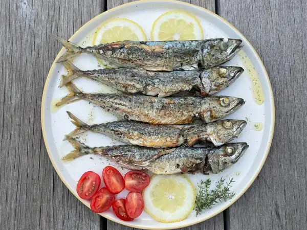 Cinq sardines au four, dorées et savoureuses, servies avec des quartiers de citron, des tomates cerises et des herbes fraîches sur une assiette blanche.