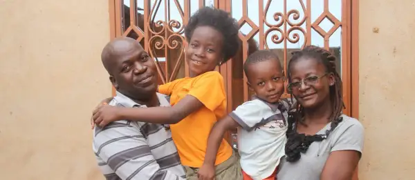 Famille souriante devant une fenêtre ornée. L'homme porte un enfant, la fille étreint son père. L'origine d'Anthony Samama est peut-être liée à cette joie familiale.