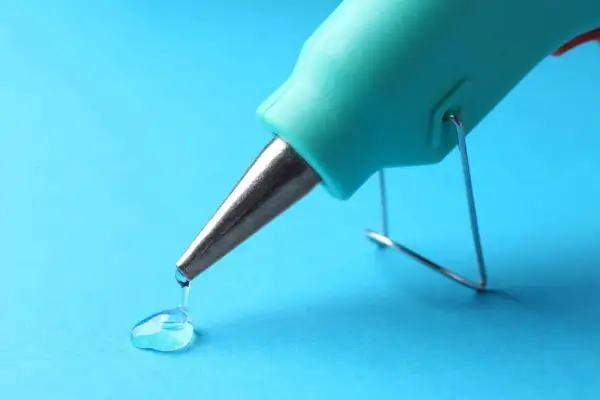 Un pistolet à colle turquoise dépose une goutte de colle transparente sur un fond bleu. Une petite tache de colle se forme.