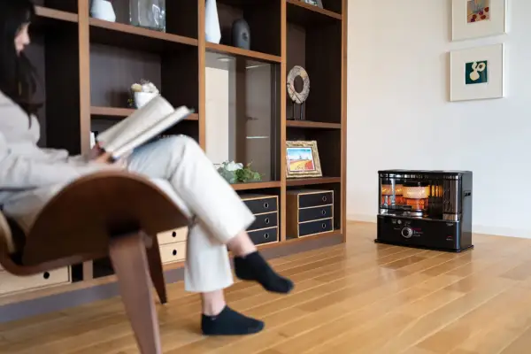 Donna legge un libro seduta su una poltrona, mentre un fungo a cherosene scalda l'ambiente. Una casa senza riscaldamento, ma con un tocco di calore.