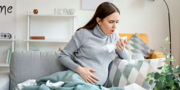 Kobieta w ciąży kaszle, szukając informacji, co na kaszel w ciąży. Na kanapie leżą chusteczki.