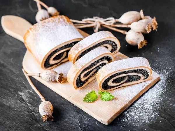 Stary przepis na makowiec, pyszny, zawinięty w ciasto i posypany cukrem pudrem. Obok suszone maki.
