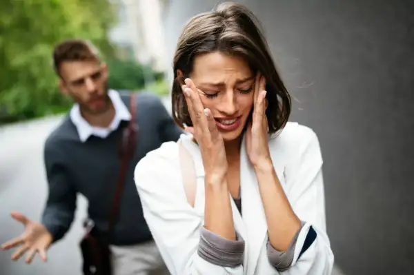 Una donna piange disperata, con le mani sul viso, mentre un uomo alle sue spalle gesticola con frustrazione. L'immagine cattura un momento di profonda infelicità di coppia.