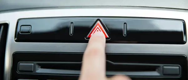 Un doigt appuie sur le bouton des feux de détresse obligatoires, un triangle rouge lumineux sur le tableau de bord d'une voiture.