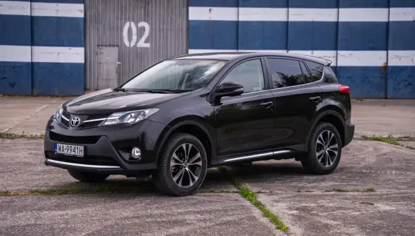 Czarna, używana Toyota RAV4 stoi na betonowym placu przed budynkiem z niebiesko-białymi pasami i numerem "02".