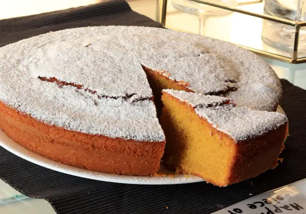 Una soffice torta carote e mandorle, cosparsa di zucchero a velo, con una fetta tagliata che ne rivela l'interno dorato.