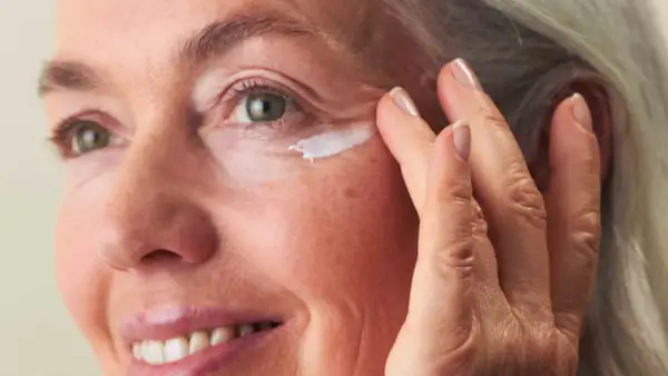 Eine Frau um die 60 trägt Augencreme auf. Ihre Haut zeigt natürliche Spuren des Alters, aber keine Pickel.