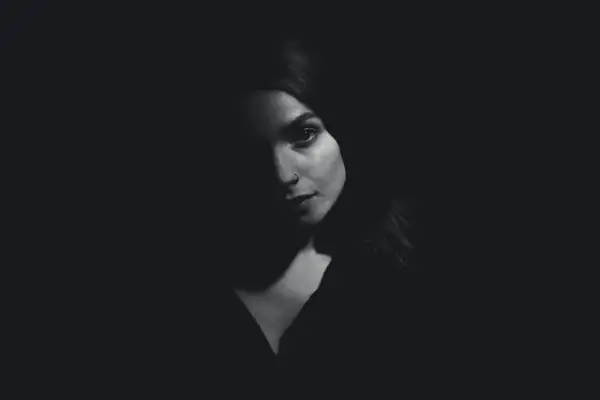 A woman's face emerges from darkness, her features dramatically sculpted by side lighting. A subtle nose ring catches the light.